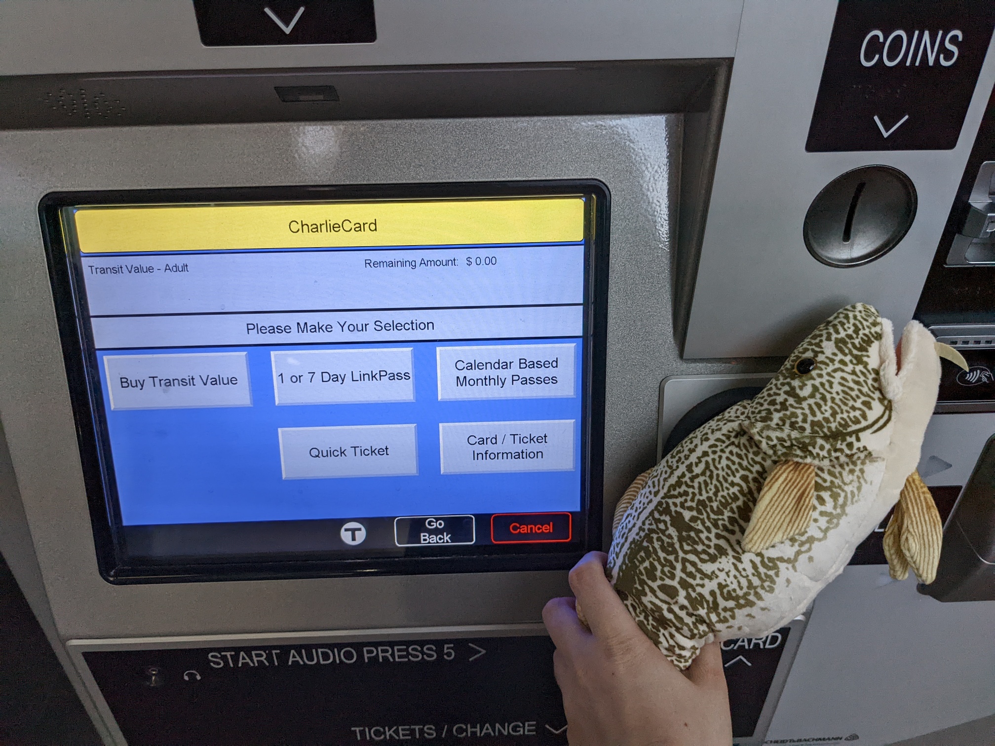 a plush codfish tapped on the reader of a MBTA ticket machine, showing a card with $0.00 on it